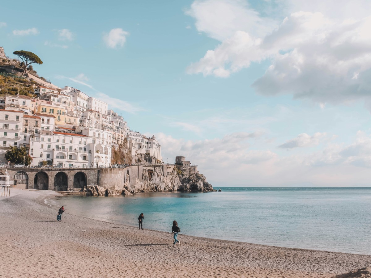 Coastal town with buildings on a cliff, sandy beach, three people, and a calm sea under a partly cloudy sky.