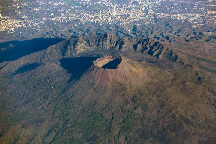 Aerial view of a large volcanic crater surrounded by rugged terrain and distant urban areas.