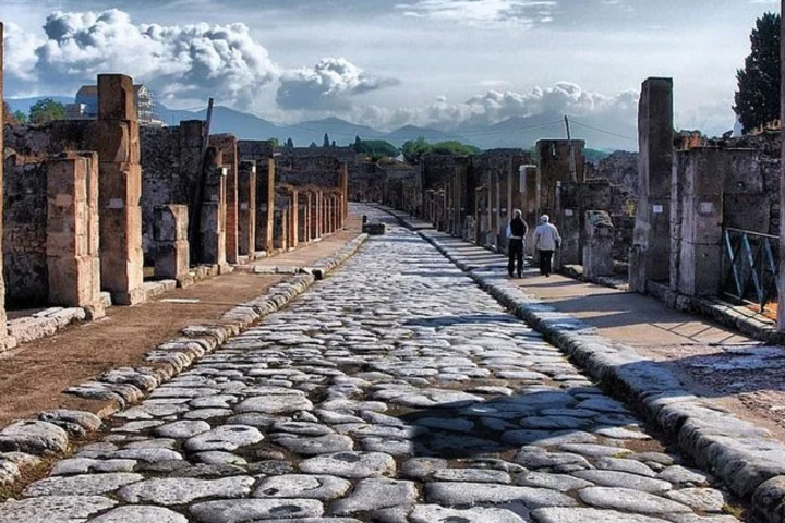 Ancient stone street with columns and two people walking, under a cloudy sky.