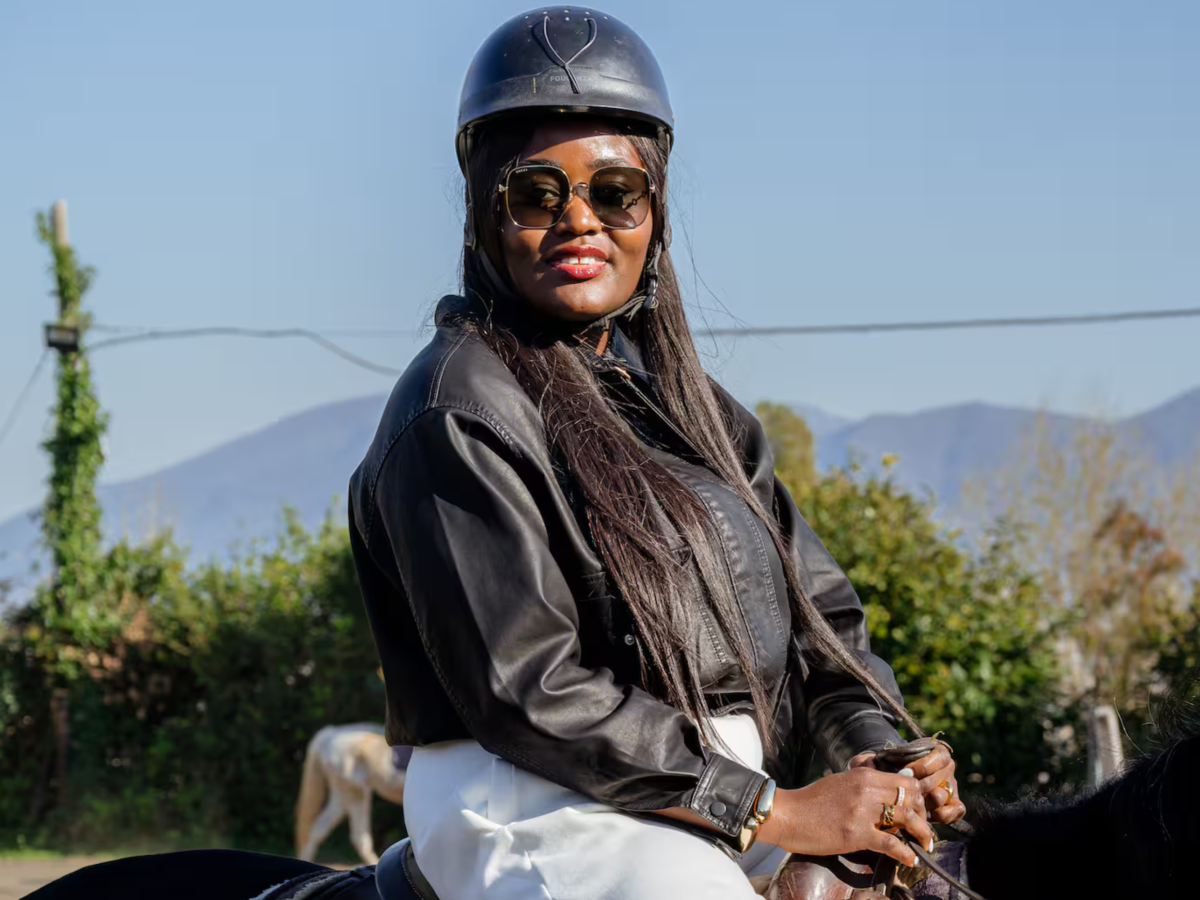 Woman wearing helmet and sunglasses, riding a horse outdoors with mountains in background.