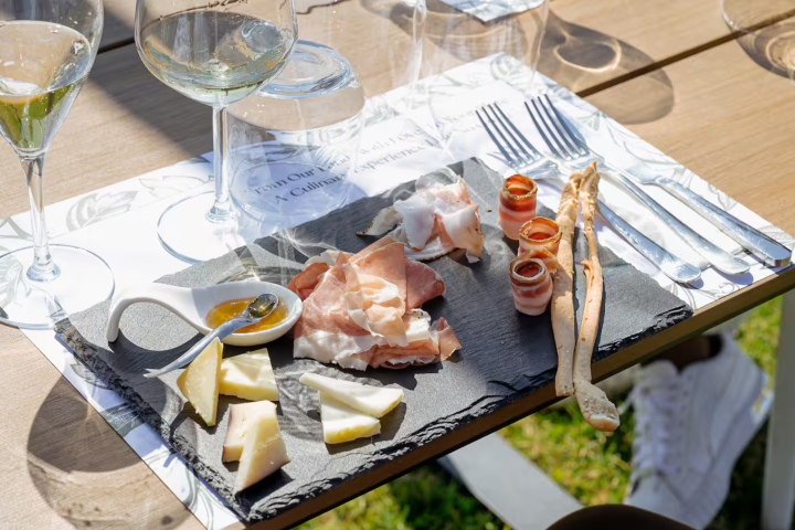 Charcuterie board with meats, cheeses, breadsticks, and sauce beside wine glasses on a wooden table.