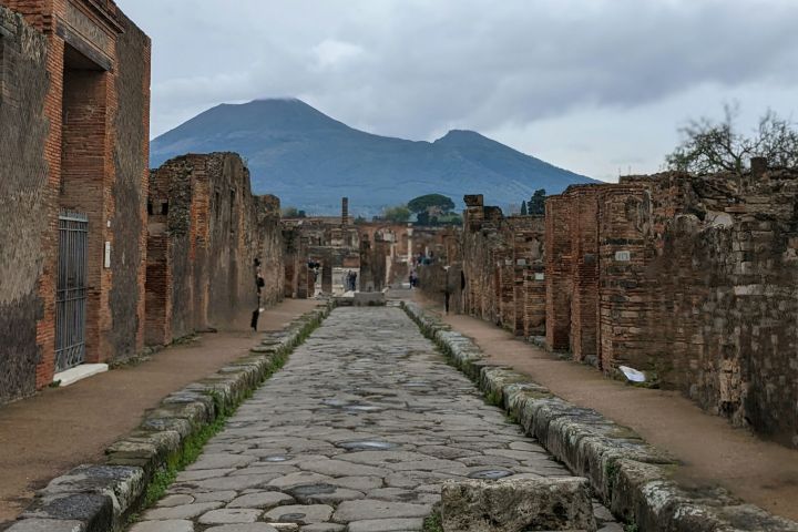 Ancient stone street with ruined buildings and mountain in background under cloudy sky.